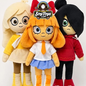 May include: Three plush dolls standing side by side. The left doll has blonde hair, a yellow shirt, and brown pants. The center doll has orange hair with a red bow, a white shirt with an orange tie, and a blue skirt. The right doll has black hair, a red hoodie, and black pants. A SoyToys.com logo is visible.