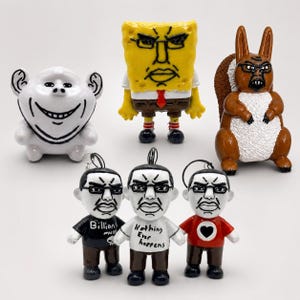 以下が含まれることがあります： 手描きされた6体の小さなフィギュアのコレクション。セットには、白いトロールのようなキャラクター、黄色いスポンジのフィギュア、茶色のリス、そして「Billions」、「Nothing Ever happens」、ハートが描かれたシャツを着た3体の人間のようなフィギュアが含まれています。