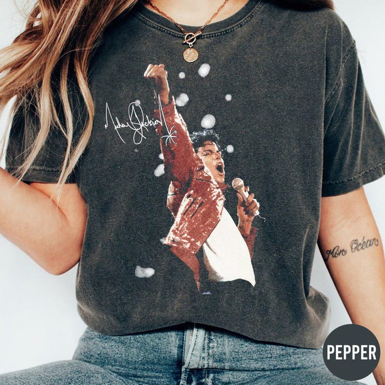 K&ouml;nnte beinhalten: Dunkelgraues T-Shirt mit einem Aufdruck von Michael Jackson in einer roten Paillettenjacke, der in ein Mikrofon singt. Das Shirt enth&auml;lt seine Unterschrift und wei&szlig;e Blasenakzente. Das Wort "PEPPER" befindet sich unten rechts.