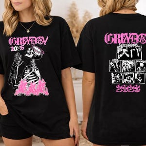 May include: Black t-shirt with a graphic design featuring a skeleton with a floral crown and pink flames. The front of the shirt has the text "GREYBOY 2025". The back of the shirt has the text "GREYBOY" and a collage of images.