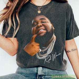 Może przedstawiać: Ciemnoszary t-shirt z krótkim rękawem z dużym nadrukiem graficznym przedstawiającym brodatego mężczyznę w srebrnych łańcuchach, wskazującego na swoje zęby. Pod portretem znajduje się napis "Rod Wave" wykonany białą kursywą.