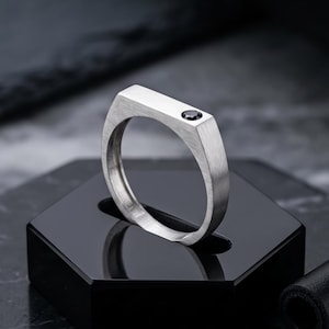 May include: A silver-toned ring with a flat, rectangular top surface featuring a single, small, round, black gemstone set flush into the metal. The band has a brushed, matte finish and a clean, geometric design with a solid, uniform structure.