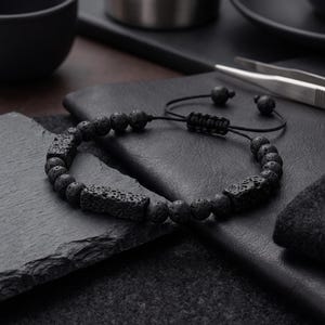 Puede incluir: Una pulsera negra con cuentas redondas de piedra volcánica porosa y dos espaciadores rectangulares de piedra volcánica texturizada. La pulsera incluye un cordón negro con cierre deslizante ajustable y dos pequeñas cuentas redondas en los extremos.
