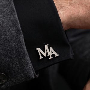 May include: A silver-toned metal cufflink featuring the letters MA in a serif font, attached to the black fabric cuff of a long-sleeved shirt worn under a dark gray textured blazer sleeve with four buttons on the cuff.