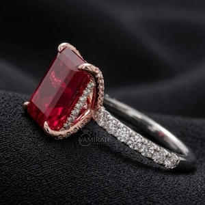 Può includere: Anello con una gemma rosso rubino a taglio rettangolare. L'anello presenta una montatura in oro rosa attorno alla pietra, impreziosita da piccole pietre trasparenti. La fascia è argentata e anch'essa incastonata con piccole pietre trasparenti.