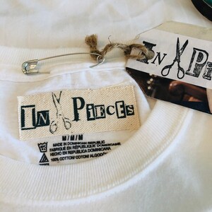 May include: Close-up of a white t-shirt with a tag that reads "In Pieces" and a safety pin. The tag also includes the text "Made in Dominican Republic" and "100% Cotton". The shirt is made of a soft, comfortable material.