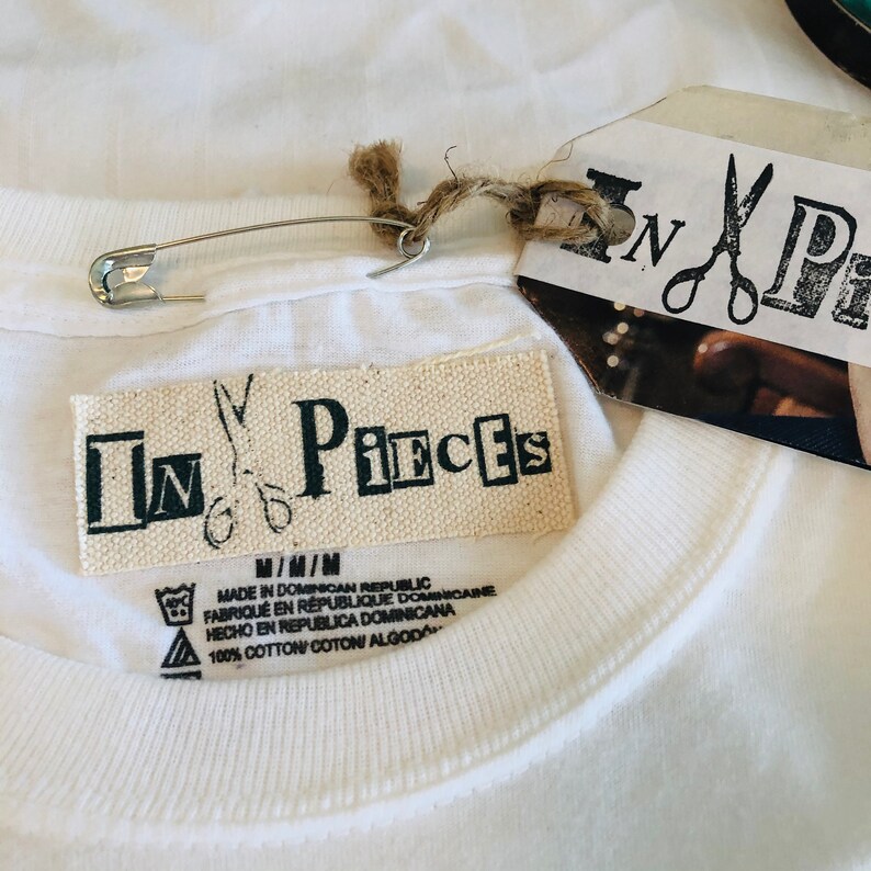 May include: Close-up of a white t-shirt with a tag that reads "In Pieces" and a safety pin. The tag also includes the text "Made in Dominican Republic" and "100% Cotton". The shirt is made of a soft, comfortable material.