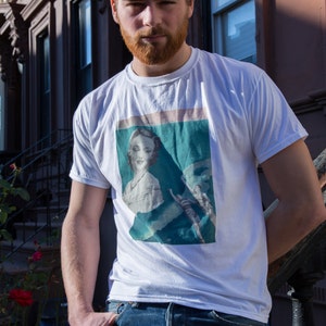 May include: White t-shirt with a vintage-style graphic print featuring a woman's portrait. The shirt is paired with blue jeans. The person is standing outdoors in front of a building with a wrought iron railing.