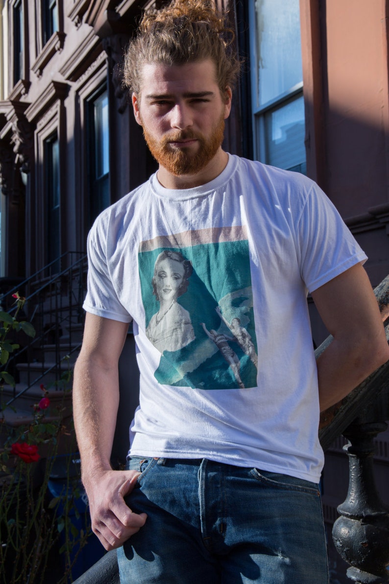 May include: White t-shirt with a vintage-style graphic print featuring a woman's portrait. The shirt is paired with blue jeans. The person is standing outdoors in front of a building with a wrought iron railing.