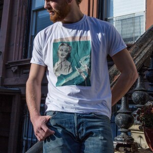May include: White t-shirt featuring a vintage-style graphic print of a woman and a figure. The shirt is paired with blue jeans. The person is standing outside near a building with a dark staircase and windows.