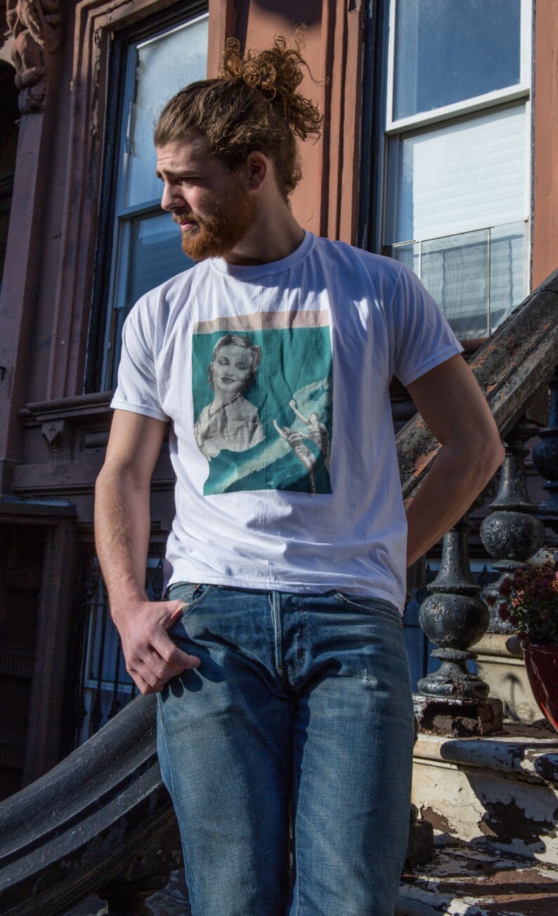 May include: White t-shirt featuring a vintage-style graphic print of a woman and a figure. The shirt is paired with blue jeans. The person is standing outside near a building with a dark staircase and windows.
