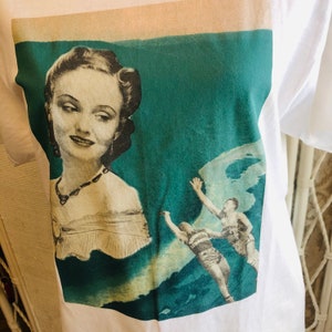 May include: White t-shirt featuring a vintage-style graphic. The graphic includes a portrait of a woman in a dress and two men in swim trunks. The background is teal and cream-colored.