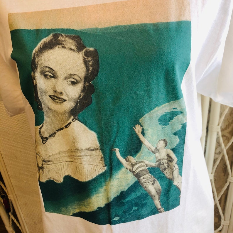 May include: White t-shirt featuring a vintage-style graphic. The graphic includes a portrait of a woman in a dress and two men in swim trunks. The background is teal and cream-colored.
