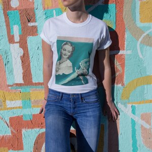 May include: White t-shirt with a vintage-style graphic of a woman in a teal setting. The shirt is paired with blue jeans with a distressed knee. The background is a colorful mural with various geometric shapes and colors.