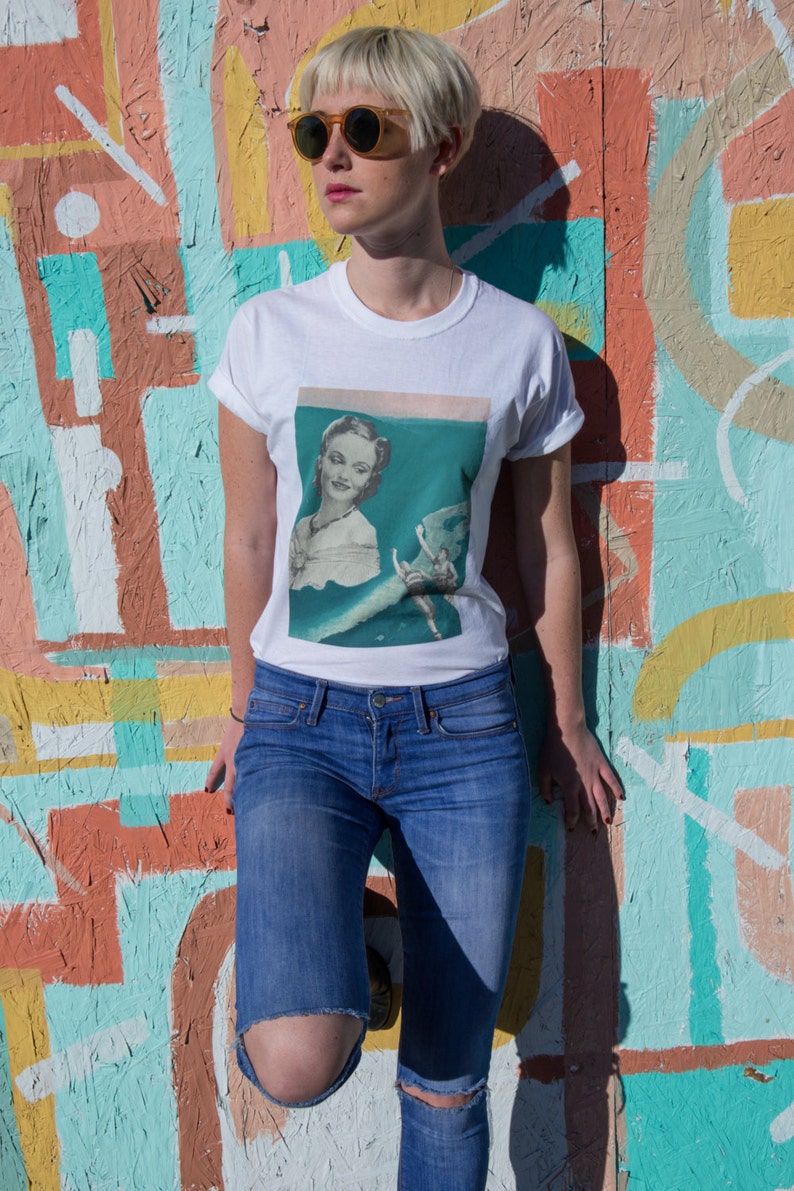 May include: White t-shirt with a vintage-style graphic of a woman in a teal setting. The shirt is paired with blue jeans with a distressed knee. The background is a colorful mural with various geometric shapes and colors.