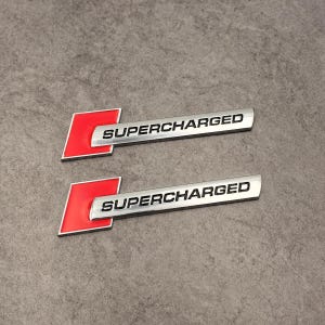 May include: Two silver-colored "SUPERCHARGED" emblems with a red and silver "C" logo. The emblems are rectangular with rounded edges and are set against a gray background.
