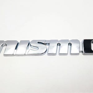 Può includere: Un'emblema cromato "NISMO" per auto con una "O" nera e cromata alla fine. L'emblema ha una superficie riflettente e un carattere audace e stilizzato. L'emblema è progettato per l'uso automobilistico.
