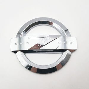 Puede incluir: Un emblema cromado brillante con un anillo exterior circular y una sección central rectangular. La sección central tiene la palabra "NISSAN" en una fuente estilizada. El emblema está sobre un fondo blanco.