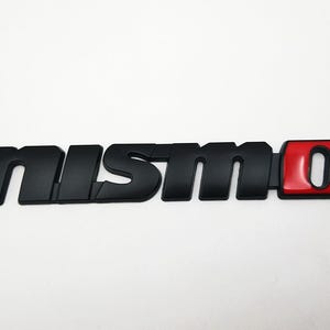May include: Black "nismo" emblem with a red and black "0" accent. The emblem is a car accessory, likely for a vehicle's exterior or interior. The letters are in a bold, sans-serif font.