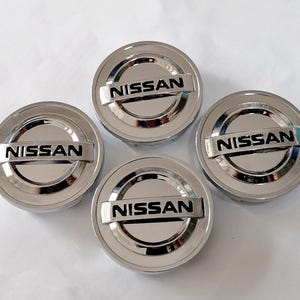 Peut inclure: Quatre enjoliveurs Nissan chromés. Chaque enjoliveur circulaire présente une section rectangulaire en relief avec le nom de la marque "NISSAN" en lettres noires. Les enjoliveurs ont un aspect métallique réfléchissant.