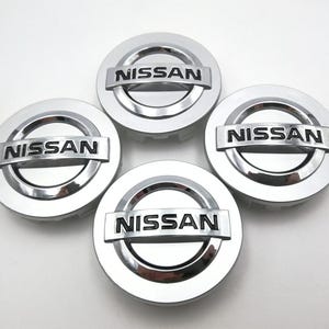 May include: Four silver Nissan hubcaps with the brand name in black lettering. The hubcaps are circular with a raised center and a shiny, reflective finish. The Nissan logo is centered on each hubcap.