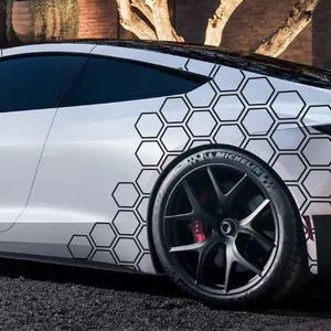 Pode incluir: Carro desportivo branco com um padrão de favo de mel preto na lateral. O carro tem rodas pretas com pinças de travão vermelhas e um pneu Michelin. O carro está estacionado numa superfície escura.