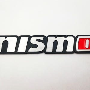 May include: A black and silver Nismo emblem with a red and silver accent. The emblem features the word "nismo" in silver, with a brushed metal texture, and a red and silver "O" at the end. The emblem is on a white background.
