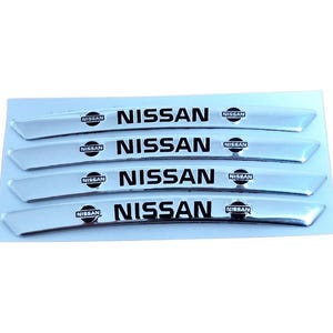 Peut inclure: Quatre autocollants d'emblème Nissan chromés. Chaque autocollant porte le logo Nissan et le mot "NISSAN" en noir, centré sur une forme rectangulaire incurvée. Les autocollants sont sur un support blanc.