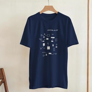Könnte beinhalten: Marineblaues T-Shirt mit weißem Grafikdesign und dem Text "LOCATION_ID: 042". Die Grafik zeigt eine Reihe verbundener geometrischer Formen. Das T-Shirt hängt an einem Holzbügel.