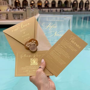 May include: Gold wedding invitation suite with calligraphy text. The suite includes an envelope, invitation, RSVP card, and details card. The envelope is sealed with a wax stamp. The invitation text includes the names Sofia and Nasser.