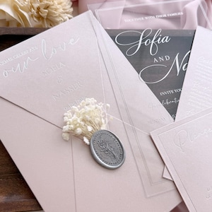 May include: A collection of wedding invitations in shades of pink and clear acrylic. The main invitation features the words "our love" and the names "Sofia & Nasser" with the date. A silver wax seal and dried flowers add detail.