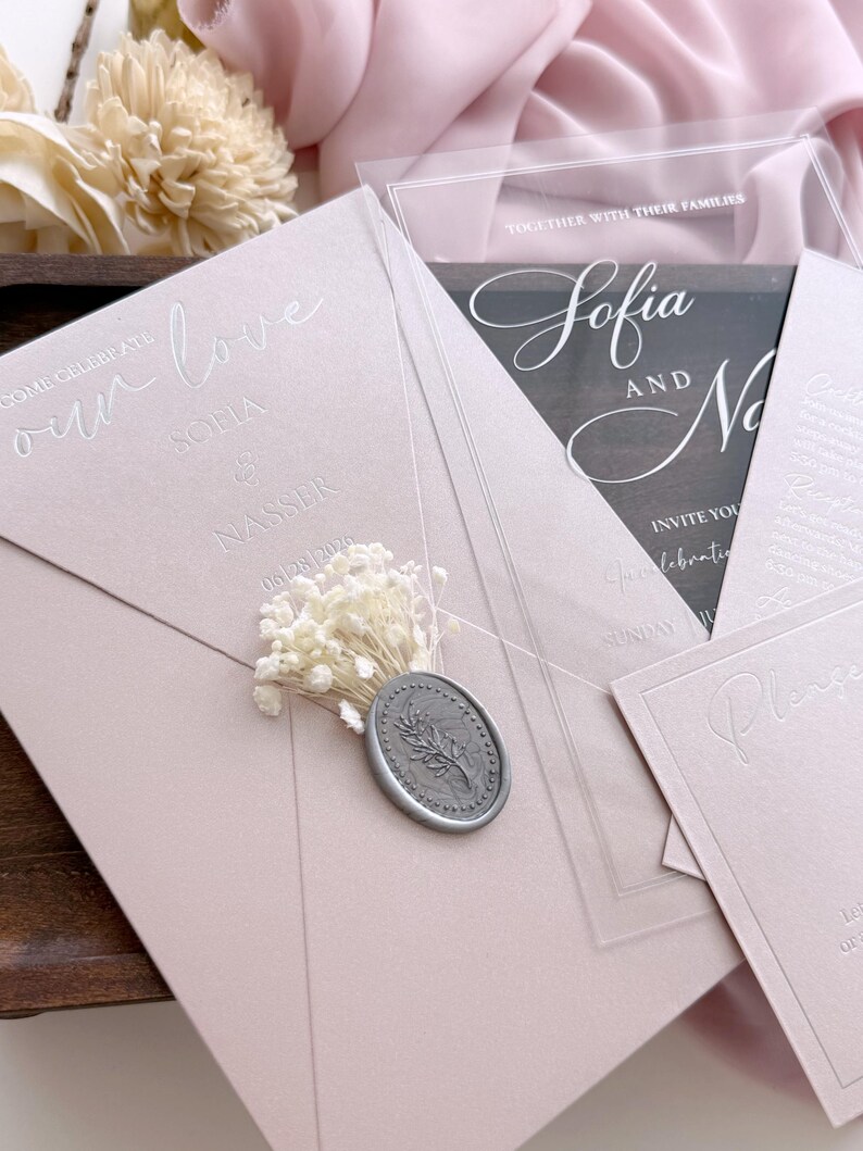 May include: A collection of wedding invitations in shades of pink and clear acrylic. The main invitation features the words "our love" and the names "Sofia & Nasser" with the date. A silver wax seal and dried flowers add detail.