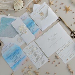 Könnte beinhalten: Ein Hochzeitspapeterie-Set mit weißen Umschlägen mit blauen Aquarell-Einlagen, einer gewölbten Einladung, einer Detailkarte und einer RSVP-Karte mit goldenem Text. Das Set enthält ein Wachssiegel, einen antiken Metallschlüssel, Muscheln und Perlen.