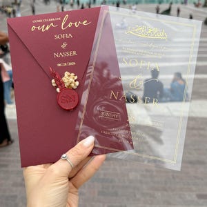 May include: Burgundy wedding invitation with gold lettering. The invitation includes the names Sofia & Nasser, date, and location. A clear overlay with gold text is held next to the invitation. The invitation is sealed with a red wax seal.
