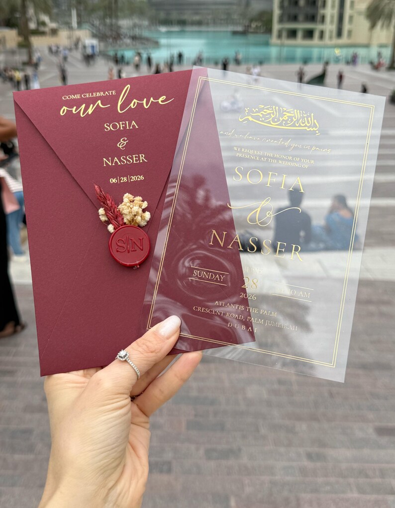 May include: Burgundy wedding invitation with gold lettering. The invitation includes the names Sofia & Nasser, date, and location. A clear overlay with gold text is held next to the invitation. The invitation is sealed with a red wax seal.