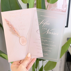 May include: A blush pink wedding invitation suite with gold foil lettering. The invitation features the names "Sofia and Nasser" and the date June 26, 2026. A dried floral element and wax seal add a decorative touch.