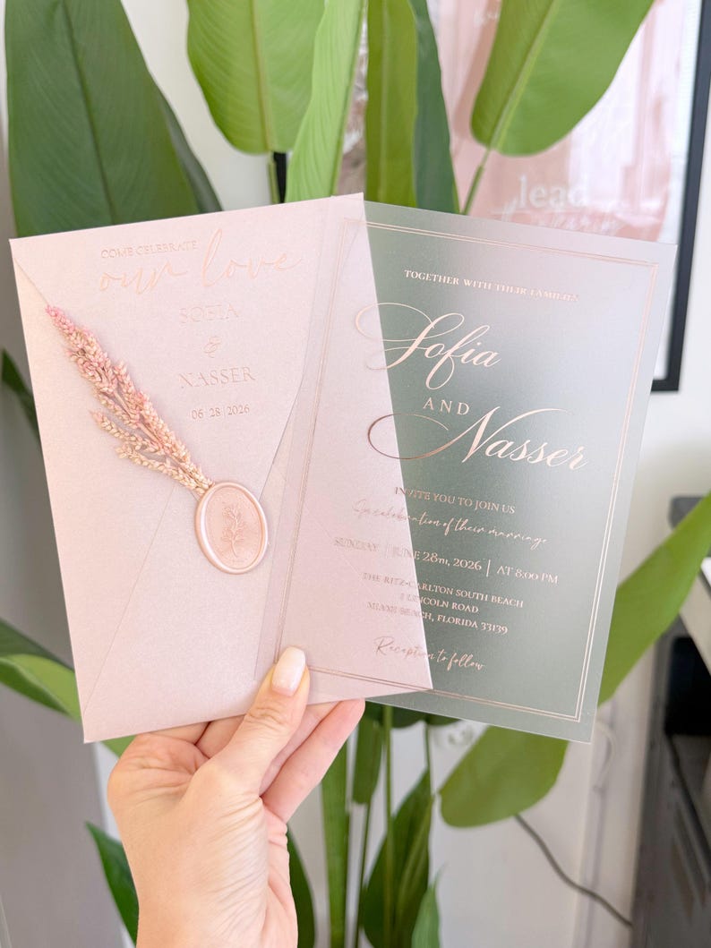 May include: A blush pink wedding invitation suite with gold foil lettering. The invitation features the names "Sofia and Nasser" and the date June 26, 2026. A dried floral element and wax seal add a decorative touch.