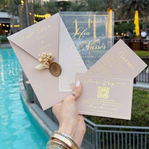 May include: A collection of wedding stationery, including an invitation, RSVP card, and details card. The invitation features the names "Sofia and Nasser" in gold script. The RSVP card includes a QR code. The stationery is a soft pink color.