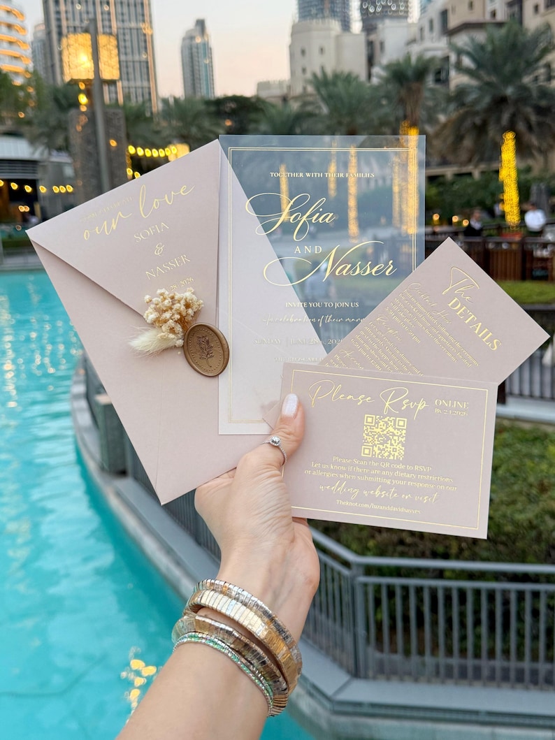 May include: A collection of wedding stationery, including an invitation, RSVP card, and details card. The invitation features the names "Sofia and Nasser" in gold script. The RSVP card includes a QR code. The stationery is a soft pink color.