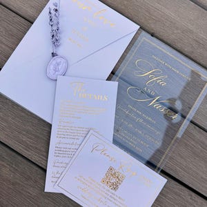 May include: A collection of wedding stationery, including an invitation, RSVP card, and details card. The invitation features gold foil lettering on a clear acrylic background. The stationery is light blue and white, with gold accents and a lavender embellishment.