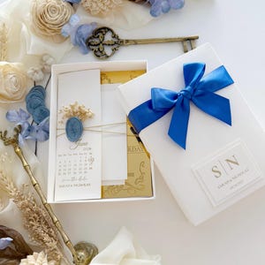 Puede incluir: Una caja blanca de invitación de boda atada con una cinta azul real. En el interior, se ve un calendario de junio de 2026, con los nombres "Sarah & Nicholas" y la fecha "09.24.2025". La invitación está adornada con flores secas y un sello de cera azul.