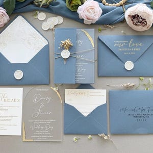 Puede incluir: Elegantes invitaciones de boda en azul empolvado con detalles dorados. El conjunto incluye sobres, tarjetas de confirmación de asistencia y tarjetas de detalles. Algunas tarjetas presentan los nombres "Daisy & Javier" y "Priscilla & Eber". Las invitaciones están selladas con sellos de cera y adornadas con flores secas.