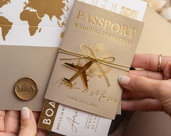 Può includere: Un invito a nozze a tema passaporto con dettagli in lamina d'oro. L'invito presenta un ciondolo a forma di aereo dorato ed è legato con uno spago dorato. Include una carta d'imbarco e una busta con una mappa del mondo.