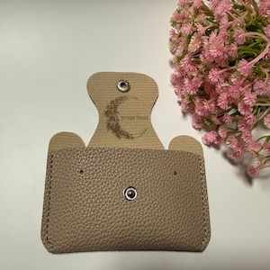 Könnte beinhalten: Ein beigefarbener Kartenhalter aus genarbtem Leder mit Druckknopfverschluss. Die Innenklappe zeigt ein eingeprägtes Mondsichel-Design mit floralen Akzenten und dem Text "your text". Der Kartenhalter hat einen rechteckigen Körper.