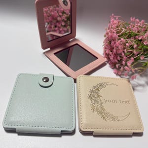 May include: Three square pocket mirrors with faux leather covers. One is open, revealing a mirror, while two are closed. The closed mirrors are light blue and beige, with the beige one featuring a crescent moon, floral design, and the text "your text".