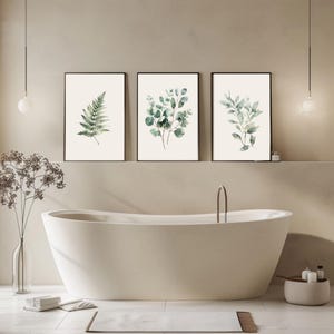 May include: Three framed botanical prints featuring watercolor illustrations of ferns and eucalyptus leaves. The prints have black frames and are displayed above a white bathtub in a neutral-toned bathroom. The artwork adds a touch of nature to the space.