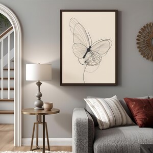 May include: A framed wall art print featuring a black line drawing of a butterfly on a cream background. The butterfly has delicate, overlapping wing outlines and a solid black body. The artwork is displayed in a dark brown frame on a gray wall.