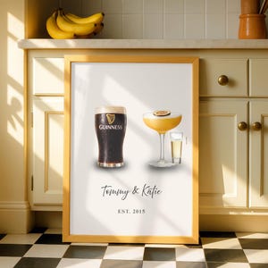 Puede incluir: Impresión enmarcada con una pinta de Guinness y un cóctel con una rodaja de maracuyá. La impresión incluye los nombres "Tommy & Katie" y el año "EST. 2015". El marco es de madera clara.