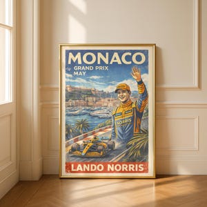 May include: A framed vintage-style poster for the Monaco Grand Prix featuring a Formula 1 race car and driver Lando Norris. The poster has a gold frame and depicts a coastal scene with the text "MONACO GRAND PRIX MAY".