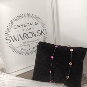 以下が含まれることがあります： 赤とゴールドのクリスタルが付いた繊細なネックレスが、黒いベルベットのクッションに展示されています。「Crystals from Swarovski Since 1895」の文字が入った透明な箱の前にネックレスが置かれています。